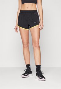 Pantalones cortos deportivos negros con ribete amarillo, cinturilla elástica y aberturas laterales. Combinados con zapatillas deportivas negras y calcetines negros con estampado.
