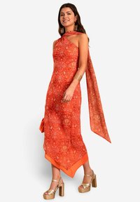 Vestido rojo con estampado, con un dobladillo asimétrico, diseño de cuello halter y detalle de larga bufanda. Combinado con sandalias de plataforma doradas con tiras.