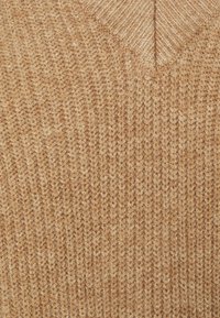 Pull en maille à col en V beige, présentant un motif tricoté texturé. Matière douce avec une teinte uniforme et chaleureuse et un détail de couture visible.