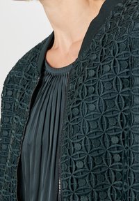 Närbild av en mörkgrön plisserad blus under en zipjacka med upphöjd blommönsterbroderi och geometriskt mönster.