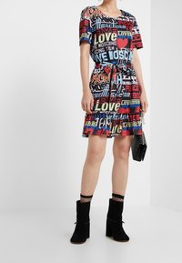 Vestido multicolorido con texto gráfico en rojo, amarillo y azul sobre un fondo negro. Mangas cortas, cintura ajustada y dobladillo por encima de la rodilla.