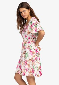 Vrouw met lang bruin haar die een korte witte bloemenjurk draagt met roze, perzik- en groene accenten, staand met haar handen achter haar rug.