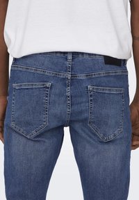 Blå denimjeans har en standard bakdesign med två fickor, subtila sömmar och en svart midjebandsetikett, sedd bakifrån.