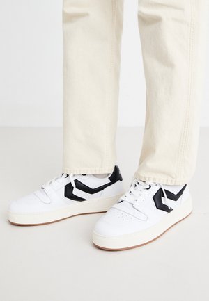 Hvide sneakers med sorte Chevron-accenter båret med cremefarvede bukser på en lys baggrund.