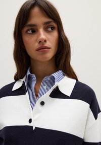 Marineblau und weiß gestreifter Pullover mit einem Kragen, der eine Knopfleiste aufweist, sowie einem darunter getragenen, kontrastierenden blau und weiß gestreiften Hemd.