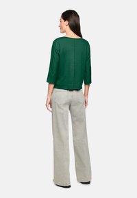 Grünes Oberteil mit drei Viertelärmeln in lässigem Schnitt, kombiniert mit hellgrauen, weit geschnittenen Hosen. Der Stoff wirkt weich und hat eine minimale Textur.