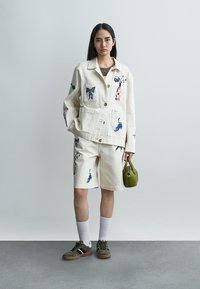 Γυναίκα που φοράει εκρού μπουφάν και σορτς με πολύχρωμα ζωικά σχέδια, λευκές κάλτσες, αθλητικά παπούτσια, κρατώντας μικρή πράσινη τσάντα, στέκεται μπροστά σε απλή τοίχο.