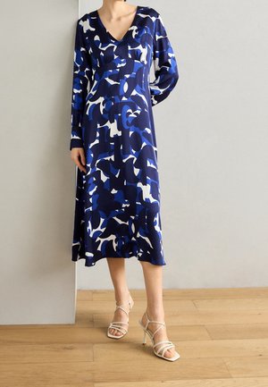 Femme portant une robe midi à motifs abstraits bleus, blancs et noirs, à manches longues, avec des sandales à talons hauts blanches à brides.