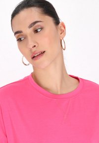 Vaaleanpunainen crewneck-huppari, jossa on hienovarainen ommeltu yksityiskohta ja sileä pinta. Mallilla on päällään kultaiset rengaskorvakorut. Tausta on yksinkertainen valkoinen.