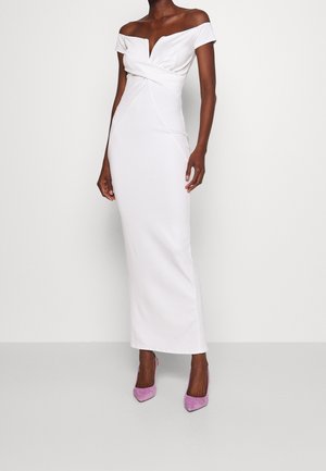 Kvinde iført en hvid gulvlængde kjole med bare skuldre og en snoet detalje ved taljen samt lyserøde spidse høje hæle, stående mod en ensfarvet baggrund.