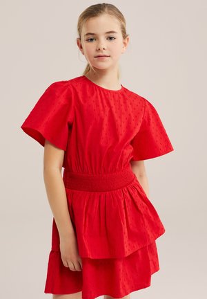Jeune fille aux cheveux blonds portant une robe rouge texturée avec des manches bouffantes et une taille smockée, debout devant un fond uni.