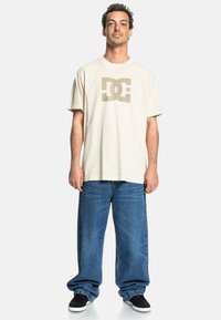 Bežové bavlněné tričko s logem DC ve světle hnědé barvě, spárované s volnými modrými džínami a černými semišovými botami.