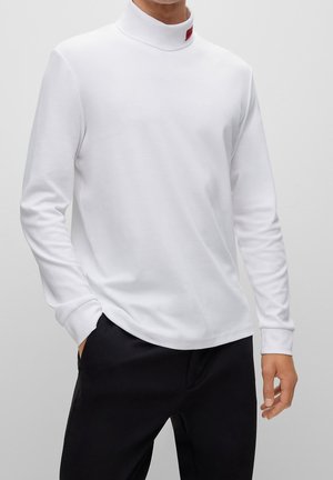 Biała koszulka z długim rękawem i golfem o wysokim kołnierzu. Gładka bawełniana tkanina. Czerwony detal metki na kołnierzu. W połączeniu z czarnymi spodniami.