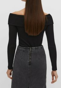 Vrouw met lang, steil bruin haar draagt een zwarte ribgebreide top met blote schouders en een donkergrijze denim rok met hoge taille en ritssluiting aan de achterkant.