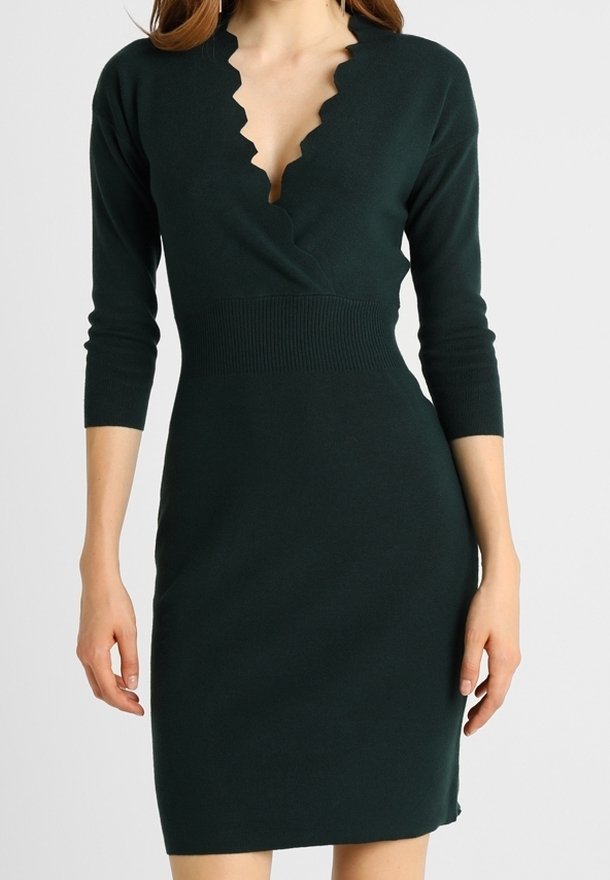 Femme porte une robe ajustée vert foncé à manches trois-quarts et encolure en V festonnée, debout devant un fond uni.