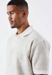 Beige poloshirt met korte mouwen, een gestructureerd patroon, een zachte, gebreide kraag en een subtiele logo in lichte borduursel op de borst.