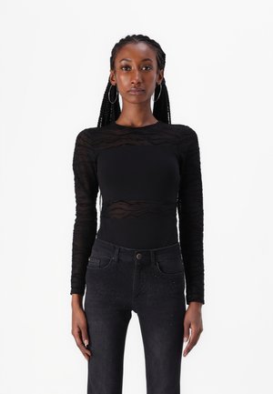 Musta pitkähihaisen paita, jossa on läpinäkyvä raidallinen tekstuuri, yhdistettynä tummiin farkkuihin, joissa on ripoteltuja niittejä etutaskuissa.