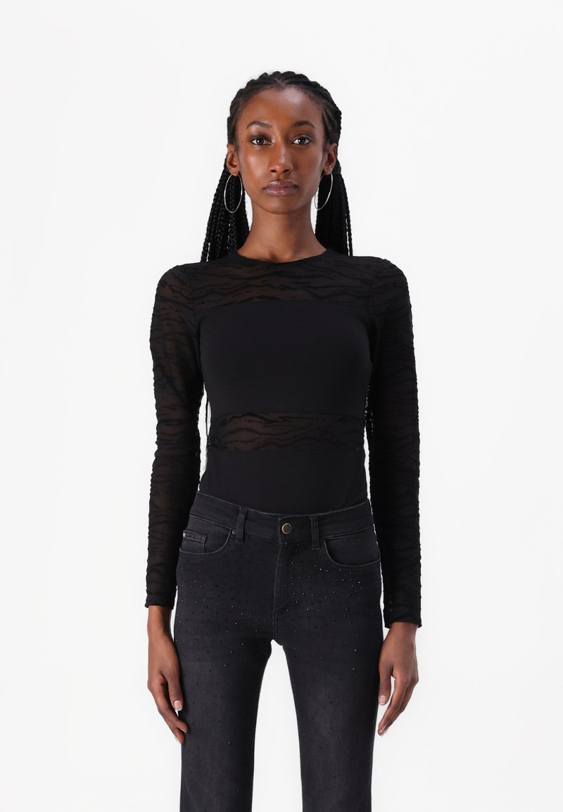 Langärmliges schwarzes Oberteil mit transparenter gestreifter Textur, kombiniert mit dunklen Jeans, die an den vorderen Taschen verstreute Nieten aufweisen.