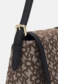 Brązowa torba z tkaniny z monogramem, czarnymi skórzanymi elementami i złotymi dodatkami. Posiada wytrzymały pasek na ramię z klamrą.