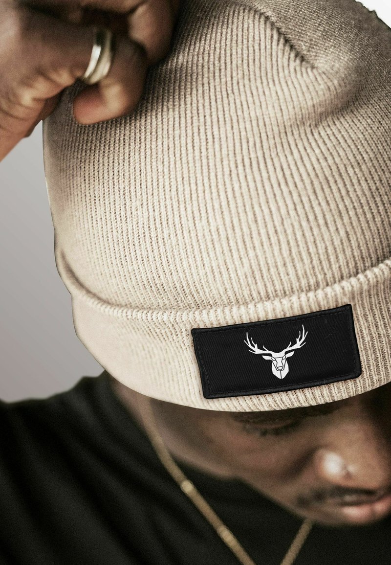 Beige knitted beanie with prominent ribbing, featuring a black rectangular patch with a white deer graphic. Soft texture, snug fit.