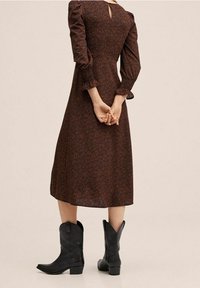 Braunes Midikleid mit Puffärmeln und Schlüsselloch-Detail am Rücken, kombiniert mit schwarzen Cowboy-Stiefeln. Das Muster besteht aus kleinen, dezenten Punkten.