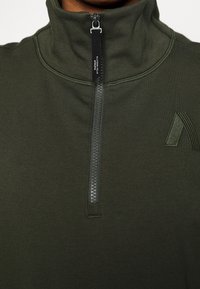 Suéter verde oscuro con un cuello alto, que presenta un cierre de media cremallera en la parte frontal y un detalle de logo en relieve en el pecho en un tono similar.