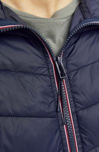 Chaqueta acolchada azul marina con cremallera negra. Presenta detalles en rojo y blanco a lo largo de la cremallera. Llevada sobre una camiseta gris de cuello redondo.