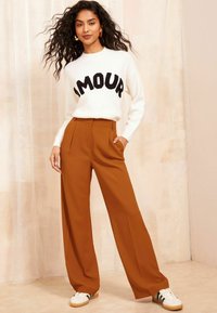 Witte trui met "AMOUR" in zwarte letters, gecombineerd met hooggetailleerde, wijde roestkleurige broeken en witte sneakers met groene accenten.