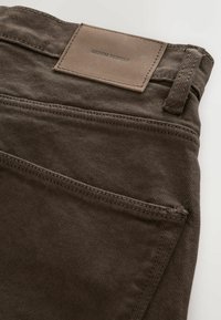 Gros plan d’un pantalon en denim marron montrant la poche arrière, la boucle de ceinture et l’étiquette en cuir indiquant "DENIM SUPPLY."
