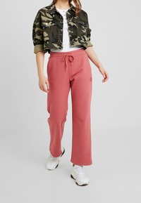 Camo cropped jack, witte henley shirt en roze sweatpants met een trekkoord. Witte sneakers maken de casual outfit compleet.