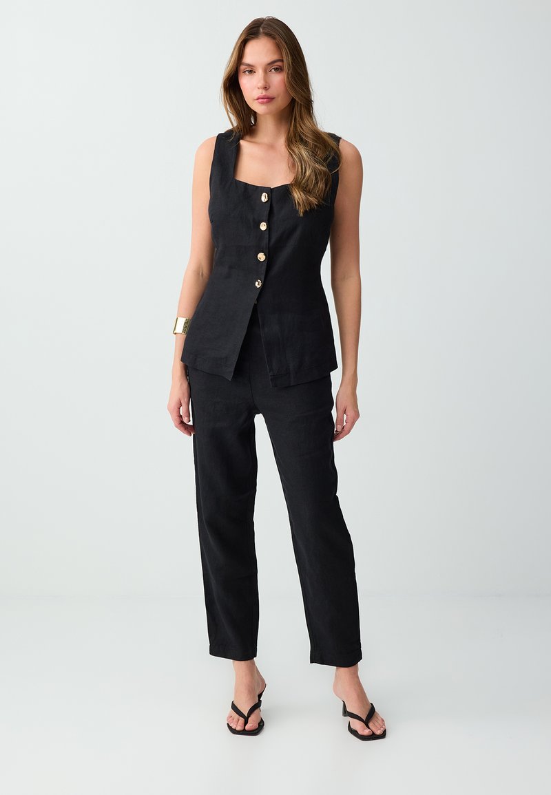 Femme portant un haut noir sans manches boutonné et un pantalon noir assorti, debout sur un sol blanc devant un fond gris clair.