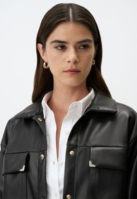 Giacca nera in ecopelle con colletto, dettagli dorati e due tasche sul petto. Indossata sopra una camicia bianca con bottoni.