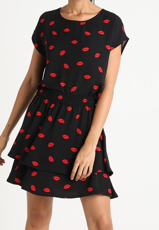 Mujer vestida con un vestido negro de manga corta y patrón de labios rojos, con falda en capas, de pie contra un fondo blanco.