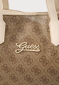 Bolso beige fabricado con material sintético texturizado con un patrón de diamantes. Presenta un destacado logo dorado de "Guess" en la parte frontal.