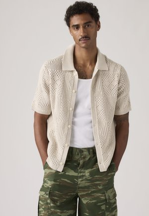 Mand iført beige kortærmet striktrøje over hvid tanktop og grøn camouflage cargobukser, stående med hænderne i lommerne.