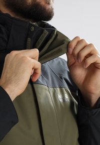 Homme à la barbe ajustant le col d'une veste Jack Wolfskin olive et noire avec fermeture éclair et fermeture Velcro.