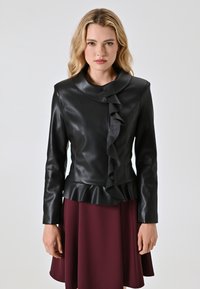 Giacca in pelle nera con dettaglio arricciato sul davanti, colletto alto e orlo peplum. Indossata sopra una gonna bordeaux, con maniche lunghe.