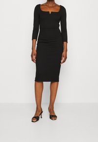 Musta vartalonmyötäinen mekko pitkillä hihoilla, neliön muotoinen pääntie jossa pieni aukko, sileä kangas, polveen ulottuva malli, yhdistettynä mustiin korkokenkiin.