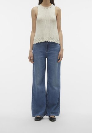 Mujer con top de punto sin mangas color crema, vaqueros azules de pierna ancha y zapatos planos negros, de pie frente a un fondo blanco liso.