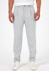 Lichtgrijze broeken met een gestructureerde afwerking, voorzien van plooien en een knoopsluiting. Gecombineerd met witte sneakers voor een casual uitstraling.