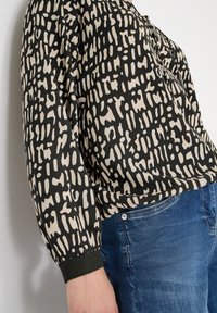 Sort og cremefarvet mønstret bluse med lange ærmer, rundet nederkant og elastiske manchetter, kombineret med blå denimjeans.