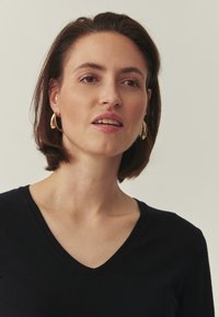 Pull noir à col en V en tissu tricoté, présentant une coupe ajustée. Le modèle porte des boucles d'oreilles créoles dorées et a des cheveux mi-longs.