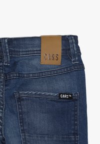 Tmavě modré džíny z denimu s hladkou texturou. Mají hnědou koženou nášivku s nápisem „CARS“ a černou značkovou visačku na zadním kapse.