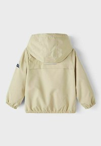 Giacca impermeabile beige con cappuccio, polsini elasticizzati e tasca posteriore a bavero. Presenta dettagli riflettenti e un logo di montagna sulla manica.
