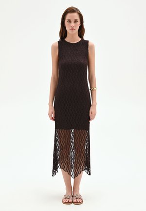 Femme portant une robe midi noire en dentelle sans manches avec un ourlet inférieur transparent, un bracelet en or au poignet gauche, et des sandales tressées beiges, debout sur fond blanc.
