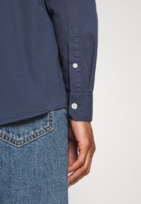 Gros plan d'une main à côté d'un jean en denim bleu et d'un poignet de chemise bleu marine à manches longues avec deux boutons blancs.