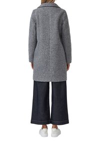 Vrouw in een lange grijze textuurjas, donkere rechttoe-rechtaan broek en witte sneakers, staand met haar gezicht naar de andere kant tegen een witte achtergrond.