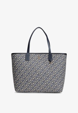Beige tote tas met marineblauw in elkaar grijpende geometrische patroon, marineblauwe handvatten en klein gouden logo-embleem aan de voorkant.