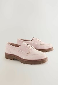 Vaaleanpunaiset suede-kengät, joissa on litteä pohja, pyöreä varvas ja koristeellinen nauhoitus. Varustettu metallipainikkeilla ja kohotetulla sauman yksityiskohdilla.