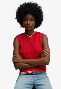 Gilet sans manches en maille rouge avec un motif torsadé, encolure arrondie, cinq boutons sur le devant et un ourlet rayé blanc et rouge.
